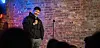 A man doing stand-up comedy in front of a brick wall.
