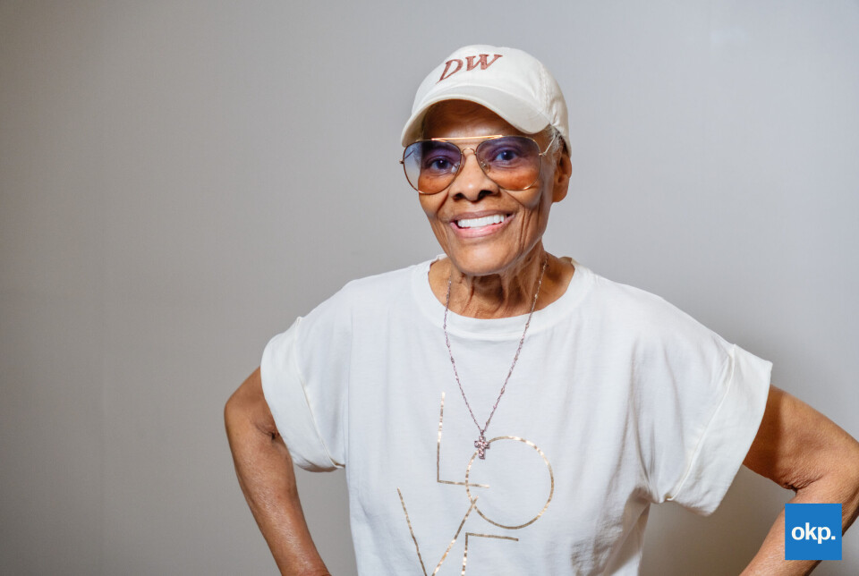 Dionne Warwick in white branded DW cap and LOVE T-shirt smiling with hands on hips