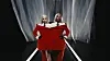 Two models stand on a lit runway sharing one large red suit before white curtains.