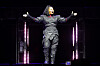 Janet Jackson performs onstage in a black outfit with arms extended, framed by vertical stage lights.