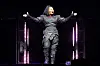 Janet Jackson performs onstage in a black outfit with arms extended, framed by vertical stage lights.