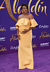 Tatyana Ali stands on a purple carpet in a strapless yellow gown at Disney’s Aladdin premiere.
