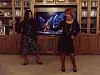 Two people standing in a living room with a large TV showing a live performance.
