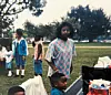 People at an outdoor park gathering with trees, grass, and picnic items.