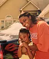 Two people smiling and posing beside a parked car in front of a house.
