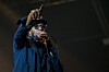 Mos Def performs onstage with a microphone, wearing a dark coat and cap under concert lighting.