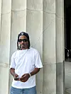 Man in a white T-shirt, sunglasses, and cap stands beside tall stone columns while holding a phone.