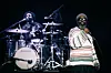 A singer stands in front of a drummer on a dark stage during a live performance.