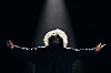 A performer in a dark outfit stands under a spotlight with arms extended on a dark stage.