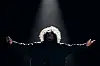 A performer in a dark outfit stands under a spotlight with arms extended on a dark stage.