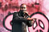 A man sings into a microphone onstage in front of a pink abstract background.