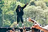 A performer on stage raises one arm above a crowd at an outdoor concert.
