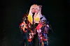 A singer in a vibrant feathered costume performs into a microphone on a dim stage.