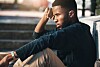 Young Black man sits outdoors in a city setting, looking tired and frustrated.