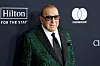Clive Davis in a green sequined jacket at a GRAMMY gala step-and-repeat with Hilton and Mastercard logos.