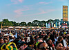 Large crowd at an outdoor festival with trees, tents and tall banners at sunset.