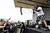 Lupe Fiasco performs on an outdoor stage while fans raise their hands in front of the barricade.