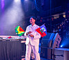 Man in a light suit holding two small flags on a stage at an indoor venue.