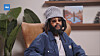 A man in a knit cap and sunglasses sits in a leather chair with shelves and plants behind him.