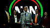 A man stands at a podium speaking, with several people behind him and a red, green and black circular backdrop.