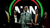 A man stands at a podium speaking, with several people behind him and a red, green and black circular backdrop.