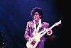Prince performing onstage with an electric guitar under purple lighting.
