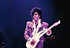 Prince performing onstage with an electric guitar under purple lighting.