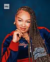 Portrait of a woman wearing a red and blue jacket with braided hair against a blue background.