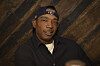 Ja Rule poses in a black shirt and cap at an indoor event with a wood-paneled backdrop.