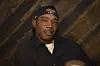 Ja Rule poses in a black shirt and cap at an indoor event with a wood-paneled backdrop.