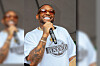 Performer in sunglasses sings into a microphone while wearing a graphic T-shirt.