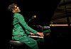 Jon Batiste in a green suit plays a grand piano on a dark stage during a CinemaCon presentation.