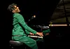 Jon Batiste in a green suit plays a grand piano on a dark stage during a CinemaCon presentation.