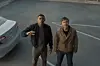 Two men look up at the camera beside a silver sedan in a parking lot.