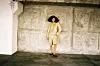 Kilo Kish wears a tan skirt and button up shirt and stands at a distance leaning against a wall.