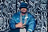 DJ Muggs poses for a photo in a blue jacket and blue hat, with a large chain.