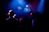 A young African American deejay performs for a crowd at a city night club. Colorful stage lights illuminate the stage behind him.