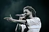 J. Cole holds a mic and points out at a crowd while wearing a white jersey.