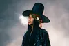 Erykah Badu wears an iconic large hat and blows smoke from her mouth on stage.