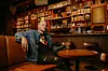 Walshy Fire sits back in a chair at an upscale bar wearing a jean jacket.