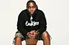 9th Wonder wearing a black hoodie, sitting down staring at the camera.