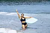 A smiling Black woman holds up a peace sign and surfboard in the water.