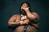 BigXThaPlug, shirtless wearing diamond jewlery, holds one hand over his face while the other grips the mic.